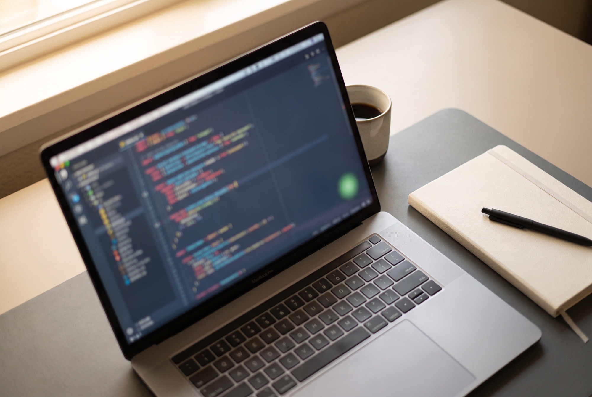 Un ordinateur portable ouvert sur un éditeur de code, un carnet et un café sur un bureau, lumière du jour.